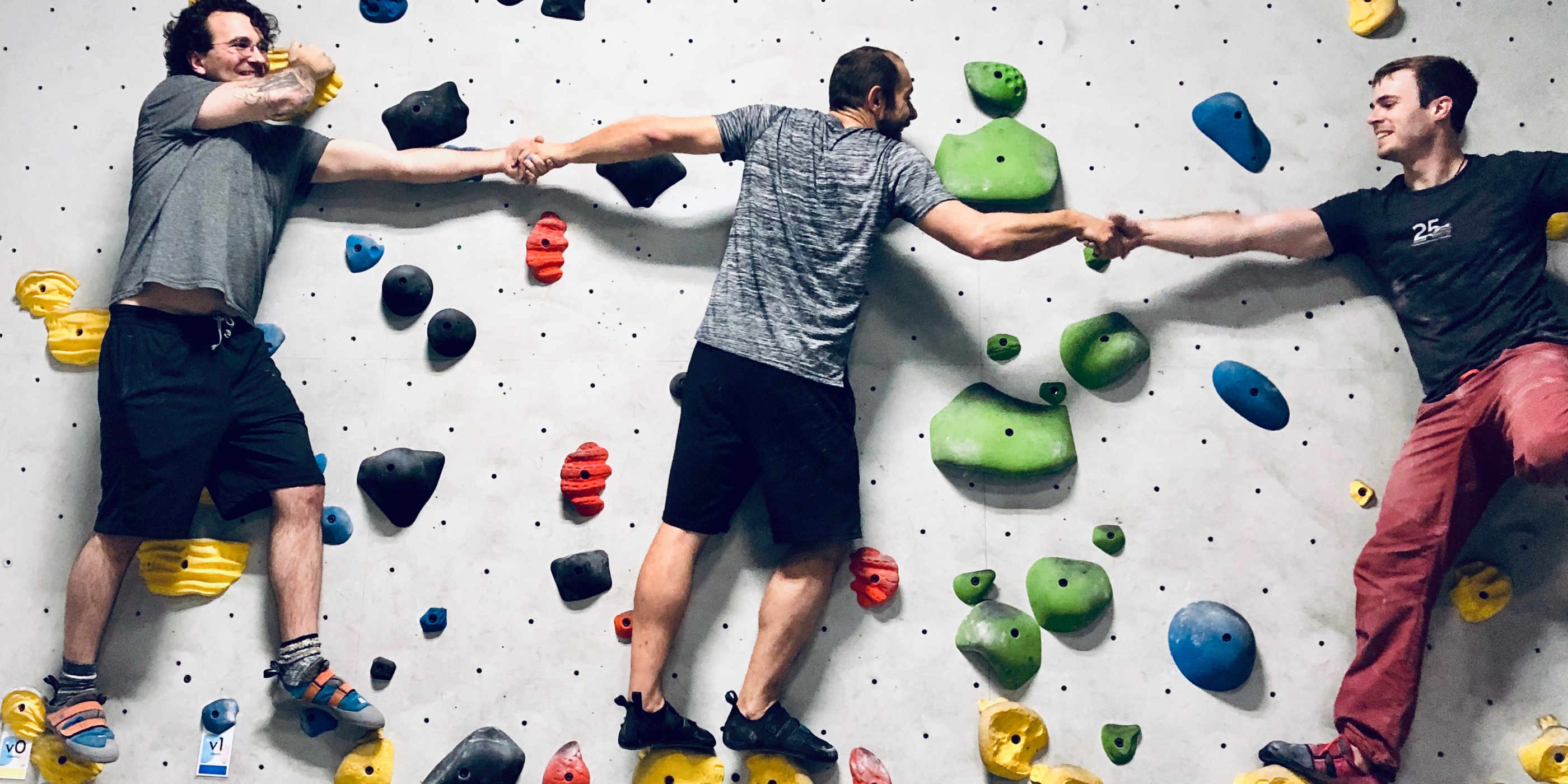 Three white guys on a rock climbing wall holding hands.