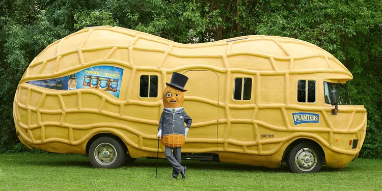 Man dressed up like a peanut with a high hat and a monocle in front of a caravan that looks like a peanut.